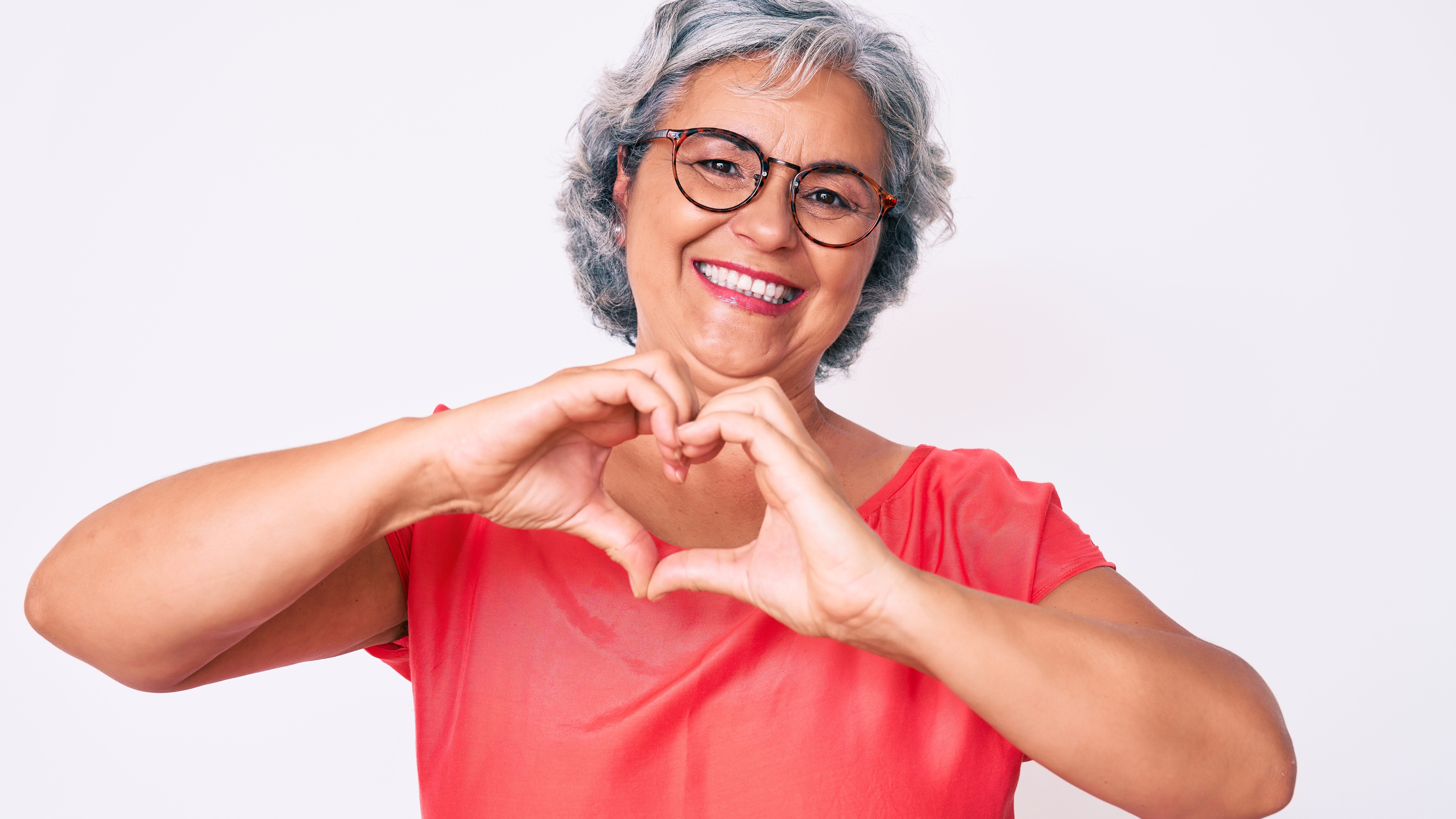 Senior hispanic grey- haired woman wearing casual clothes and glasses smiling in love doing heart symbol shape with hands. romantic concept. AdobeStock 364379527 edited - Senior hispanic grey- haired woman wearing casual clothes and glasses smiling in love doing heart symbol shape with hands. romantic concept.